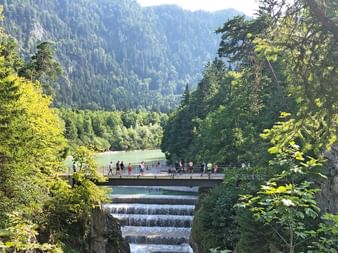 Lechfall Wasserfall mit kaskadierendem Wasser, Aussichtsbrücke mit Besuchern, eingerahmt von grünen Bäumen und bewaldeten Bergen im Hintergrund.