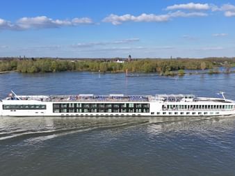 White river cruise ship MS Lisabelle sailing on blue water with forested shoreline and blue sky with white clouds in background.
