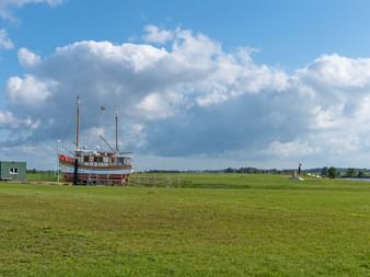 Traditional wooden sailing vessel with two masts displayed on green grass field in Greetsiel. Blue sky with white clouds and trees visible in background.