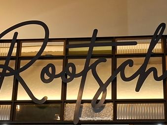 Schwarzes Schreibschrift-Schild ROOTSCH auf beleuchteten Glaspaneelen mit dekorativen Mustern in warmem goldenen Licht.