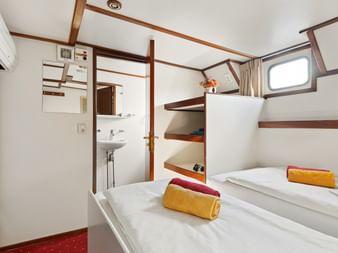 Kompakte Schiffskabine mit zwei Betten, weißen Wänden mit Holzverkleidung, Bullauge, Waschbecken, Klimaanlage und bunten Handtüchern.