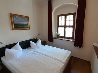 Helles Hotelzimmer mit Doppelbett, weißer Bettwäsche, Fensternische mit dunklen Vorhängen, Landschaftsgemälde und Holzboden.