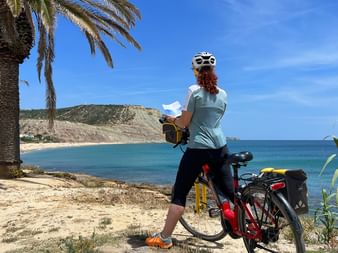 Radfahrerin mit Helm liest Karte neben Tourenrad an malerischer portugiesischer Bucht mit Palmen, türkisem Wasser und Küstenklippen.
