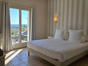 Modern hotel room with white double bed, striped accent wall, balcony doors with Provence landscape view, tile flooring, and warm wall lighting.