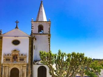 Weiße portugiesische Kirche mit steinernem Glockenturm und Kreuz auf der Kuppel vor blauem Himmel. Beschnittene Bäume und traditionelle Gebäude in der Nähe.