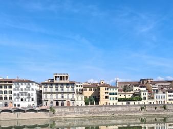 Reihe historischer italienischer Gebäude spiegelt sich im Arno in der Toskana, mit einem Kirchturm rechts unter blauem Himmel.