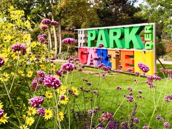 Buntes Schild mit der Aufschrift 'PARK DER GÄRTEN' in grünen, roten, blauen, orangen und gelben Buchstaben, umgeben von gelben und lila Blumen.