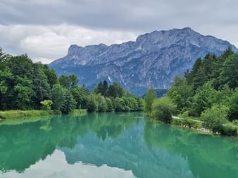 Türkisfarbener Fluss fließt durch grünen Wald mit dramatischen Berggipfeln im Hintergrund unter bewölktem Himmel in der Alpe Adria Region.