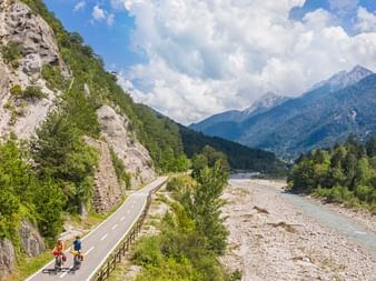 Zwei Radfahrer auf asphaltiertem Radweg entlang Flusstal mit Kalksteinfelsen, Wald und Alpenbergen unter bewölktem Himmel in Slowenien.