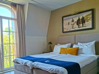 Helles Hotelzimmer mit Doppelbett, blauem Läufer, Balkontür mit Gartenblick, Strandkunstwerk und Nachttischlampen im Hotel Mondragon.