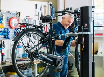 Lächelnder Fahrradmechaniker im blauen Hemd arbeitet an blauem Fahrrad im Montageständer. Werkstatt hat organisierte Werkzeugaufbewahrung.