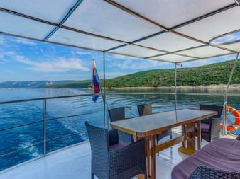 Überdachtes Deck der MY Linda mit Esstisch, Korbstühlen und lila Kissen mit Blick auf ruhiges blaues Wasser und grüne kroatische Küste.