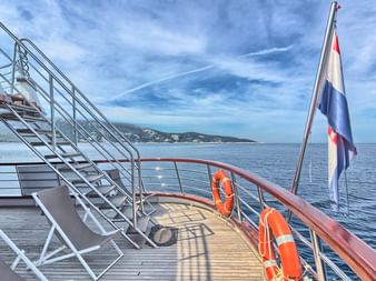Heckdeck der MY Andela Lora mit Holzboden, Metalltreppe, orangen Rettungsringen und kroatischer Flagge. Küstenhügel über ruhiger See sichtbar.