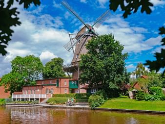 Traditional windmill with white sails beside a canal in Hinte, East Frisia. Red brick buildings and green trees surround the mill under a blue sky.