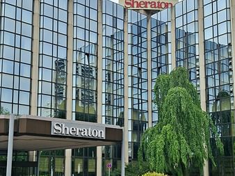 Moderne Glasfassade des Sheraton Hotels Essen mit Eingangsvordach und Garten mit geschnittenen Büschen und einer Trauerweide.