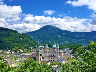 Panoramablick auf Salzburg mit barocker Kathedrale und grünen Kupferkuppeln im Vordergrund, umgeben von Alpenbergen unter blauem Himmel.