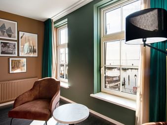 Modernes Hotelzimmer mit grünen Wänden, braunem Sessel, Stehlampe und Fensterblick. Gerahmte maritime Kunstwerke an brauner Wand.