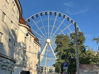 Weißes Riesenrad vor blauem Himmel in Dresden, mit historischem cremefarbenem Gebäude links und Straße mit parkenden Autos im Vordergrund.