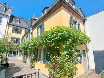 Gelbes Eckhaus mit grünen Fensterläden und weinbewachsener Pergola in Bonner Innenhof. Kopfsteinpflasterweg führt zwischen historischen Gebäuden.
