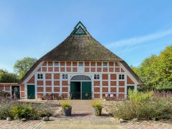 Traditionelles Fachwerkhaus mit Reetdach in Mooriem. Rot-weiße Fassade mit grünen Türen, umgeben von blühenden Gärten unter blauem Himmel.