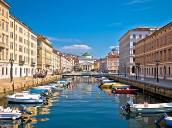 Canal Grande in Triest mit bunten Booten an Steinkai vertäut. Historische neoklassizistische Gebäude säumen beide Seiten unter blauem Himmel.