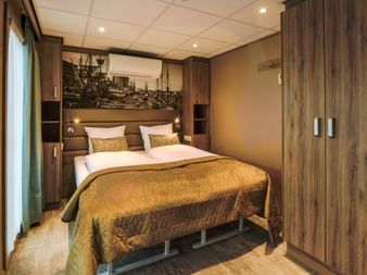 Modern ship cabin with double bed, golden bedding, wood paneling, maritime artwork above headboard, and built-in wardrobe.
