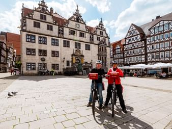 Zwei Radfahrer in roten Jacken mit Rädern auf dem Marktplatz in Hann. Münden, mit historischem Rathaus und Fachwerkhäusern.