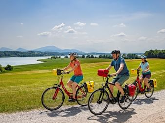 Three cyclists with touring bikes and panniers on gravel path through green fields. Lake and mountains visible in background under blue sky.