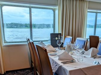 Eleganter Restauranttisch auf der SE Manon mit weißer Tischdecke, Weingläsern und Servietten. Große Fenster zeigen Flussblick mit Ufer.