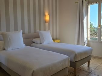 Twin bedroom with two single beds, striped beige wall, wall lamps, yellow nightstands, and window overlooking Provence countryside.