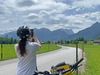Radfahrer mit Helm fotografiert Alpenpanorama. Fahrrad mit gelber Tasche im Vordergrund, Straße führt durch grüne Wiesen zu Bergen.