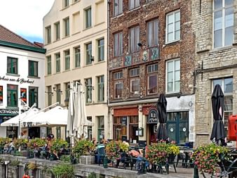 Reihe historischer Gebäude entlang eines Ufers in Brügge mit Außenterrassen, Blumenkästen und traditioneller flämischer Architektur.