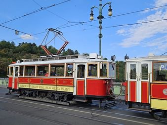 Rot-weiße historische Straßenbahn auf Gleisen in Prag unter blauem Himmel. Die Bahn zeigt den Text 'Elektrické dráhy král. hlav. města Prahy'.