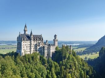 Schloss Neuschwanstein thront auf einem bewaldeten Hügel mit Türmen, umgeben von grünen Tälern und Bergen unter blauem Himmel.