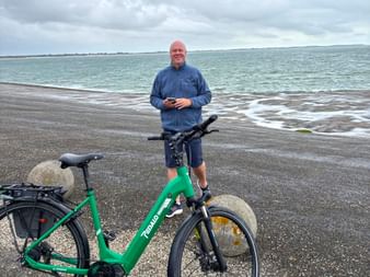 Mann in blauer Jacke mit grünem E-Bike auf nasser Küstenstraße in Neeltje Jans, Zeeland. Bewölkter Himmel und raue See im Hintergrund.
