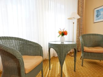 Helle Hotelsitzecke mit zwei grünen Korbstühlen mit beigen Kissen, rundem Glastisch mit Blumenvase, Stehlampe und Wandbild.