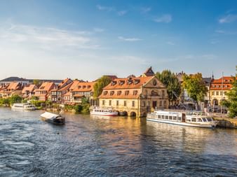Historische Bamberger Uferpromenade mit bunten Fachwerkhäusern entlang des Mains. Ausflugsboote liegen unter blauem Himmel am Anleger.