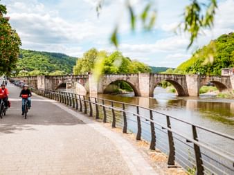 Zwei Radfahrer auf dem Weserweg in Hann. Münden mit historischer Steinbogenbrücke und grünen Hügeln im Hintergrund.