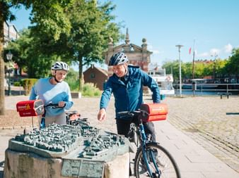 Zwei Radfahrer mit Helmen betrachten ein detailliertes Bronze-Stadtmodell von Emden auf einem Kopfsteinpflasterplatz mit historischen Gebäuden.