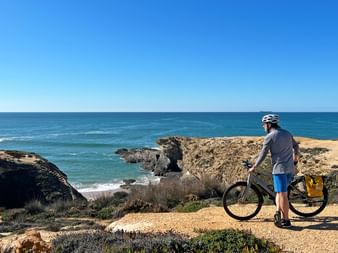 Radfahrer mit Helm steht neben Fahrrad auf Küstenklippe mit Blick auf Atlantik, felsige Küste und klaren blauen Himmel.
