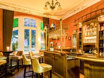 Elegante Hotelbar mit orangefarbenen Wänden, goldenem Holztresen, Messingzapfhähnen, Samtstühlen und verzierter Decke. Große Fenster mit Vorhängen.