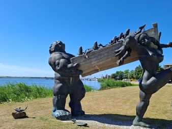 Bronzeskulptur von zwei Figuren, die ein Boot mit Menschen in Kuressaare, Saaremaa tragen. Uferpromenade und grüner Rasen im Hintergrund.
