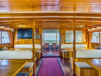 Innenraum des MY Linda Schiffsrestaurants mit Holzwänden und -decke, weißen Sitzbänken, bordeauxrotem Teppichläufer und Glastüren zum Deck.