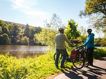 Zwei Radfahrer mit Helmen stehen mit ihrem Tourenrad auf einem Weg neben der Weser im Weserbergland, umgeben von grüner Vegetation.