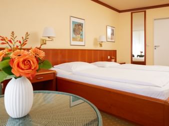 Helles Hotelzimmer mit Doppelbett, Holzmöbeln, weißer Vase mit orangen Rosen auf Glastisch und cremefarbenen Wänden.