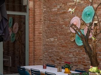 Außenterrasse mit weißem Tisch, bunten Stühlen und dekorativen Papierfächern an einem Baum vor einer Backsteinmauer in Toulouse.