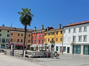 Sonniger Platz in Palmanova mit bunten Gebäuden in Weiß, Gelb, Rot und Orange. Eine hohe Palme steht neben einem Brunnen, wo Radfahrer rasten.