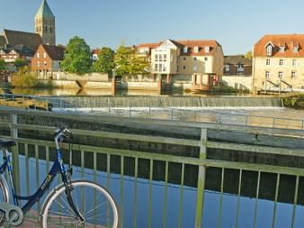 Blaues Fahrrad am Brückengeländer über der Ems in Rheine mit historischen Gebäuden und Kirchturm im Hintergrund unter klarem Himmel.