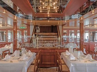 Elegantes Restaurant an Bord der MS Switzerland mit weiß gedeckten Tischen, Holzstühlen und einem zentralen Klavier. Warme Beleuchtung und Fenster.