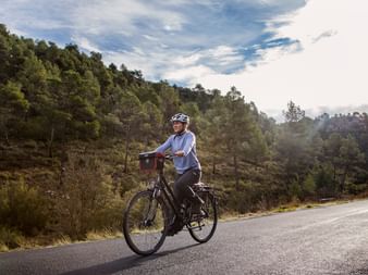 Radfahrerin mit Helm fährt auf einer Asphaltstraße durch bewaldete Hügel in der Region Valencia in Spanien unter teilweise bewölktem Himmel.